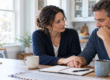 Two adults sitting together at a kitchen table, one offering a supportive hand during a difficult conversation.