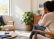 Woman sitting in a sunlit living room, resting her chin on her hand and looking out the window in quiet thought