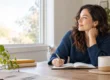 Adult writing in a notebook at a table while reflecting by a bright window.