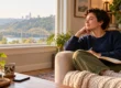 Adult sitting by a sunlit window with a notebook and phone, reflecting in a calm home setting.